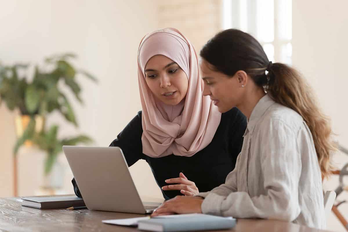 Concentrated young arabian woman in hijab sitting with smiling colleague at table, looking at computer screen, explaining new company software. Focused team leader training millennial female intern.
