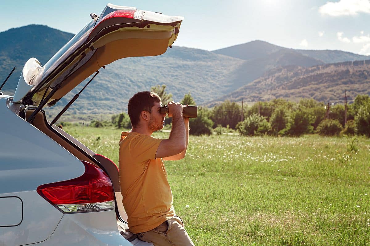 man at mountain looking through binoculars sitting on the trunk of a car; more inclusive screening process concept