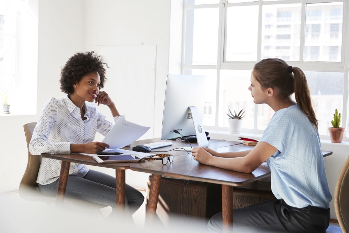 Two young women talking across their desks in an office