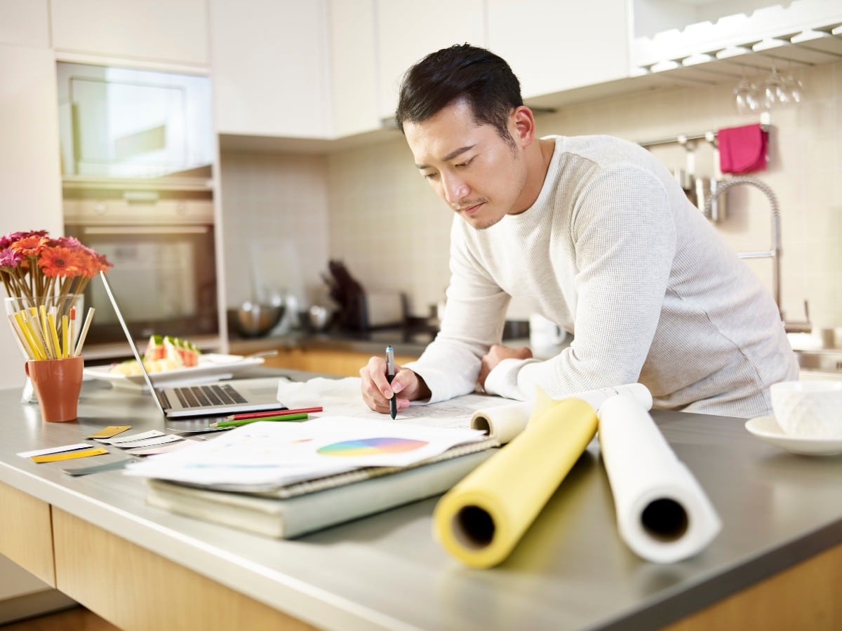 man with laptop and stacks of charts work from kitchen counter; remote compensation, localization, remote pay concept