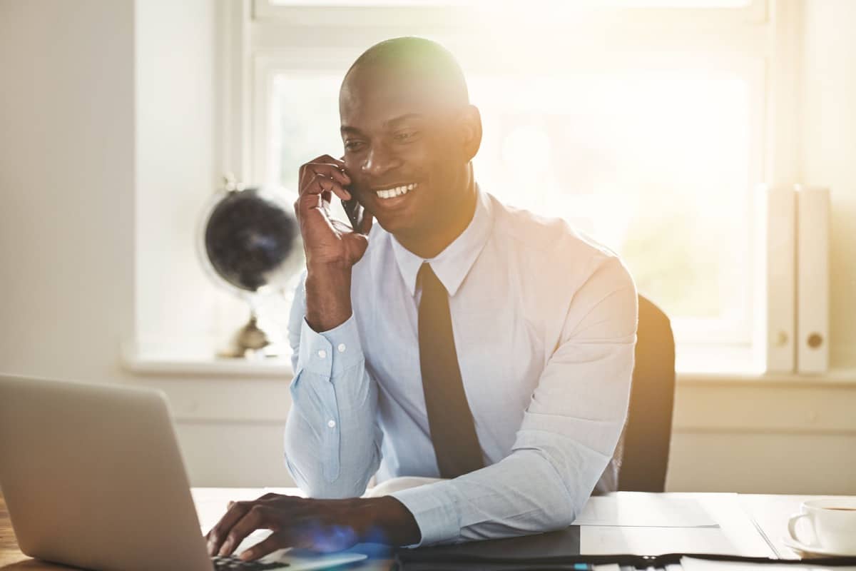 Smiling young African businessman talking to a client on his cellphone while sitting at his desk in an office
