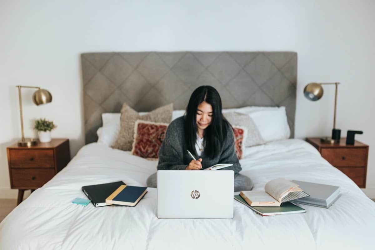 woman working from home on her bed, with laptop and papers strewn about; post-pandemic remote work concept