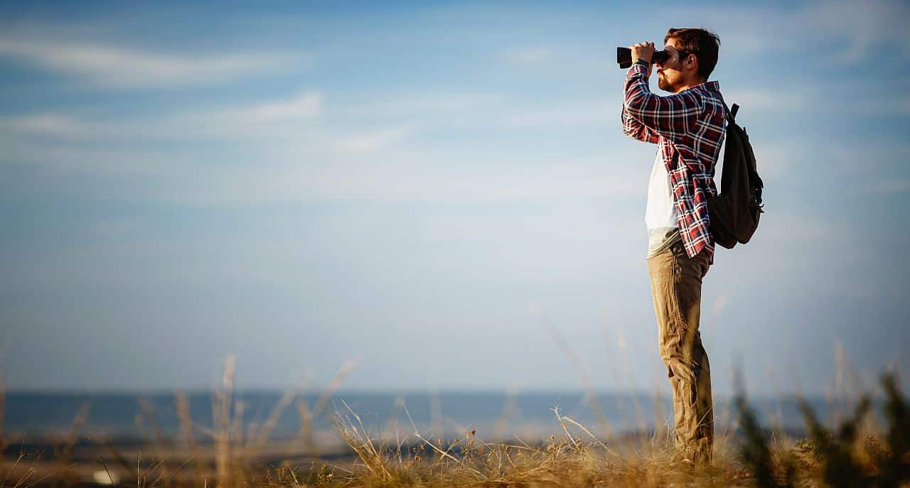 Guy looking at binoculars in hill. man in t-shirt with backpack. Young Caucasian man during hike in valley landscape