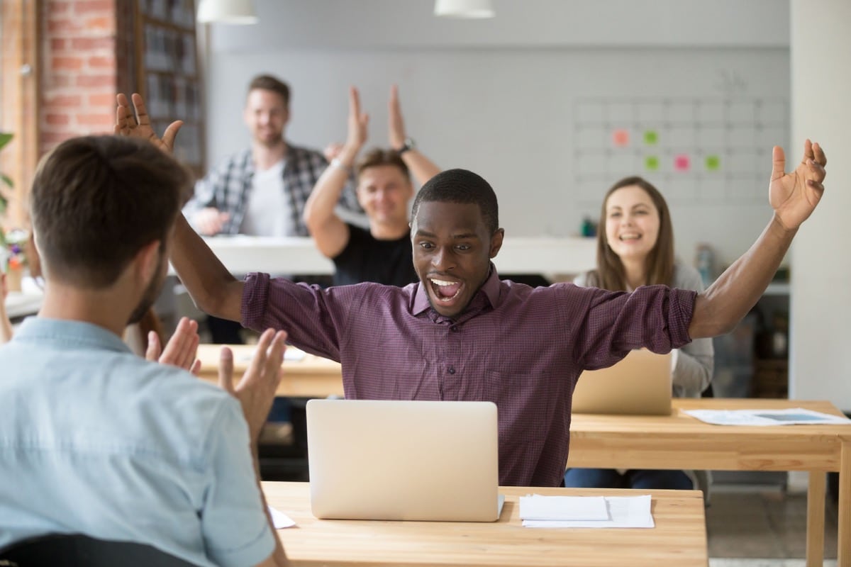 Employee excited about win, raising hands looking at laptop, coworking office team applauding; overcoming imposter syndrome concept