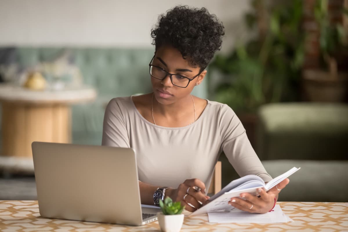 Focused young african american businesswoman looking at laptop holding book; reskilling employees concept